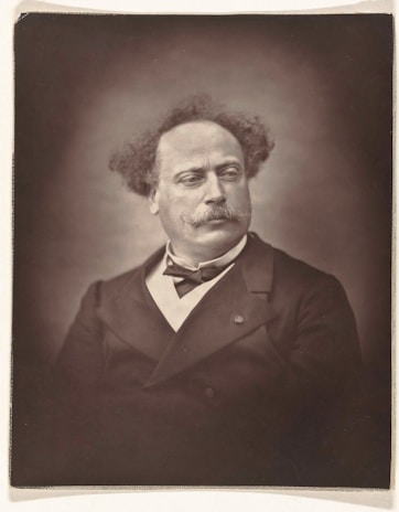 A sepia-toned portrait of a middle-aged man with distinct curly hair and a prominent mustache. He is wearing a dark suit with a bow tie, and his expression is thoughtful and slightly contemplative. The background is softly blurred, focusing attention on the subject's facial features and attire.