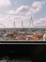 A cityscape view showcasing numerous buildings with red-tiled roofs and some notable architectural landmarks outlined and labeled, including a cathedral with a prominent spire. Below the city view, there is a dark interior with a tray and a glass placed on a ledge.