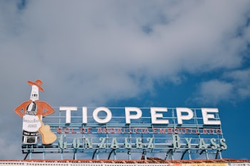 A neon sign depicting a bottle man character with a red jacket and hat holding a guitar, above the text 'TIO PEPE' in large letters. Below, the words 'Sol de Andalucía embotellado' and 'Gonzalez Byass' are displayed. The background features a blue sky with scattered clouds.
