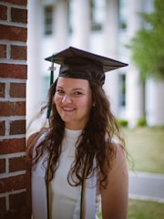 A young woman with long curly hair is standing next to a brick wall. She is wearing a graduation cap and gown, with a tassel indicating the year 2021. The background shows an out-of-focus building and some greenery.