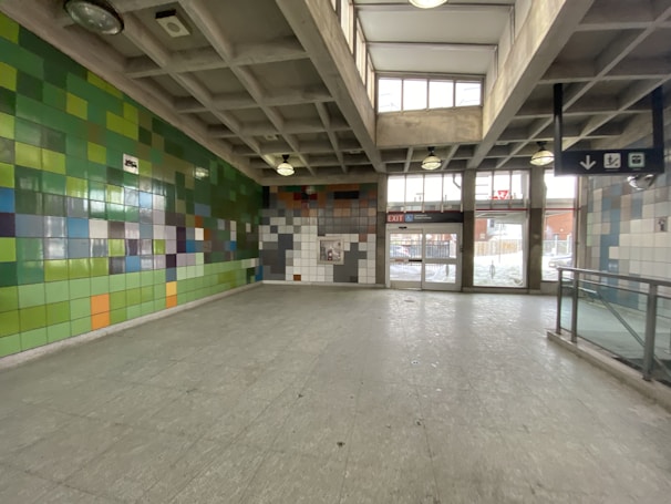 A spacious indoor area with colored tiled walls in shades of green, blue, orange, and gray. The ceiling is constructed with concrete beams and several circular pendant lights. Large windows above the entrance allow natural light to illuminate the space. A sign indicating an exit and directional icons are visible.