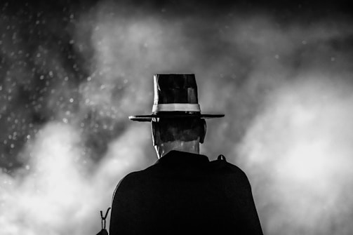 A black and white image depicting a person from behind, wearing a wide-brimmed hat and a dark cloak. The background is filled with smoky mist, creating an eerie and mysterious atmosphere.