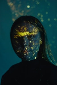 A person with long hair is illuminated by a pattern of light projections that resemble stars or galaxies against a dark background. The projections create a mystical and otherworldly atmosphere as they highlight parts of the face with glowing spots.