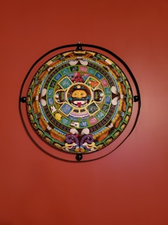 A vibrant and intricately designed circular artwork featuring a variety of colorful symbols and figures. The center displays a face surrounded by geometric shapes and detailed patterns. The artwork is mounted on a vibrant red wall, adding contrast to its colorful design.