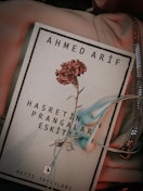 A book cover featuring the name Ahmed Arif at the top and a red carnation in the center. Below the flower, there is text in Turkish. A pair of light blue earphones is placed on the book, which rests on a soft fabric with a zippered section visible.