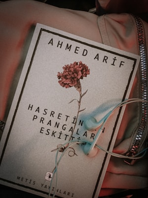 A book cover featuring the name Ahmed Arif at the top and a red carnation in the center. Below the flower, there is text in Turkish. A pair of light blue earphones is placed on the book, which rests on a soft fabric with a zippered section visible.