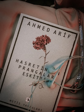 A book cover featuring the name Ahmed Arif at the top and a red carnation in the center. Below the flower, there is text in Turkish. A pair of light blue earphones is placed on the book, which rests on a soft fabric with a zippered section visible.
