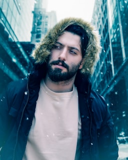 A man with a beard wearing a fur-lined hooded jacket stands on a snowy city street. Tall buildings with glass facades surround him. Snowflakes gently fall, creating a cold and wintry atmosphere.