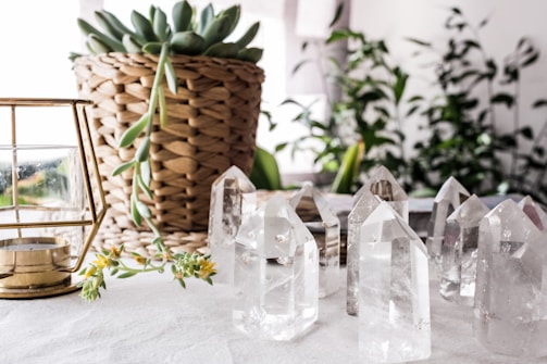 A collection of clear crystal points arranged on a white surface next to a potted plant with a woven basket texture. A brass and glass candle holder sits on the left, and there are additional plants visible in the blurred background.