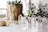 A collection of clear crystal points arranged on a white surface next to a potted plant with a woven basket texture. A brass and glass candle holder sits on the left, and there are additional plants visible in the blurred background.