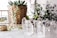 A collection of clear crystal points arranged on a white surface next to a potted plant with a woven basket texture. A brass and glass candle holder sits on the left, and there are additional plants visible in the blurred background.