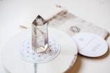 A clear quartz crystal stands upright on a circular, decorative mandala-patterned base. The background includes a soft fabric pouch with a geometric design and a round card with text, adding to the aesthetic appeal.