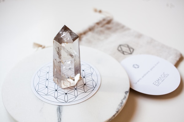 A clear quartz crystal stands upright on a circular, decorative mandala-patterned base. The background includes a soft fabric pouch with a geometric design and a round card with text, adding to the aesthetic appeal.