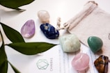 Various polished gemstones in different colors are arranged on a light surface, accompanied by a small fabric pouch and partially covered text. Green leaves from a plant partially frame the scene, adding a natural element.