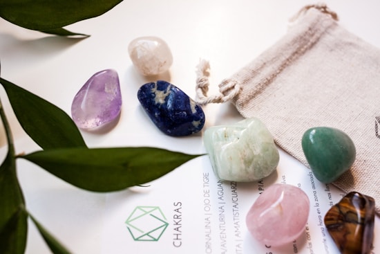 Various polished gemstones in different colors are arranged on a light surface, accompanied by a small fabric pouch and partially covered text. Green leaves from a plant partially frame the scene, adding a natural element.