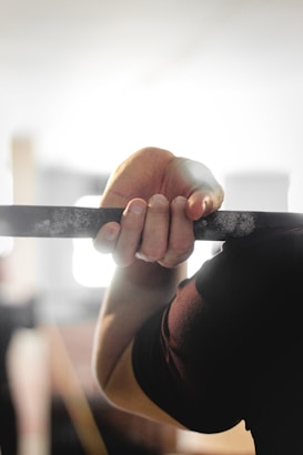 A close-up view of a person's hand gripping a bar, likely a pull-up bar. The grip is firm and the background is softly blurred, suggesting an indoor gym or workout environment.