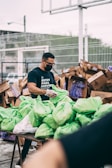 A man wearing a black shirt and face mask is organizing and handling green plastic bags on a table. In the background, there are several large cardboard boxes and a fenced area. The man appears to be involved in some form of volunteer or community service work.