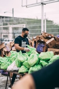 A man wearing a black shirt and face mask is organizing and handling green plastic bags on a table. In the background, there are several large cardboard boxes and a fenced area. The man appears to be involved in some form of volunteer or community service work.