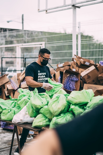 A man wearing a black shirt and face mask is organizing and handling green plastic bags on a table. In the background, there are several large cardboard boxes and a fenced area. The man appears to be involved in some form of volunteer or community service work.