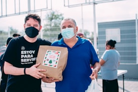 Two individuals, both wearing masks, are holding a paper bag with a label that reads 'Entrega Especial' in Spanish, indicating a special delivery. The person on the left is wearing a black shirt that says 'Estoy Para Servir' which translates to 'I am here to serve'. People are present in the background, possibly engaged in an outdoor community or volunteer activity.