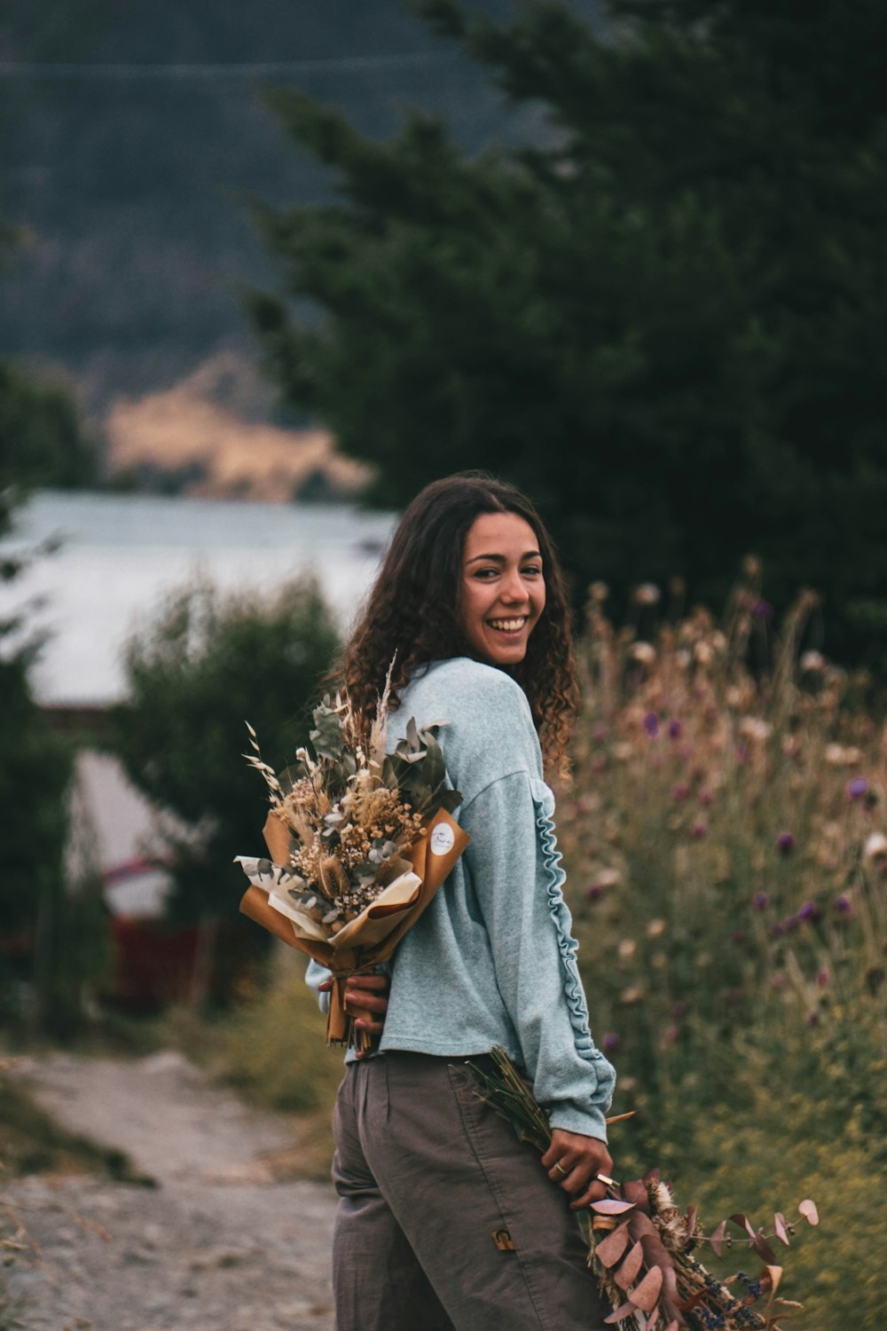 a woman walking down a dirt road holding a bouquet of flowers