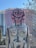 A large, abstract sculpture depicting a humanoid figure with exaggerated features, including large ears and prominent facial markings. The sculpture has a rustic, weathered appearance with muted colors, and it stands outdoors against a backdrop of trees and a partially obscured building under construction.