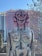 A large, abstract sculpture depicting a humanoid figure with exaggerated features, including large ears and prominent facial markings. The sculpture has a rustic, weathered appearance with muted colors, and it stands outdoors against a backdrop of trees and a partially obscured building under construction.