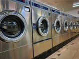 A row of industrial washing machines is lined up in a laundromat. The machines are metallic with glass doors, reflecting light from overhead fluorescent lighting. The floor consists of large tiles with visible wear.