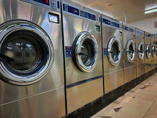 A row of industrial washing machines is lined up in a laundromat. The machines are metallic with glass doors, reflecting light from overhead fluorescent lighting. The floor consists of large tiles with visible wear.