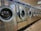 A row of industrial washing machines is lined up in a laundromat. The machines are metallic with glass doors, reflecting light from overhead fluorescent lighting. The floor consists of large tiles with visible wear.
