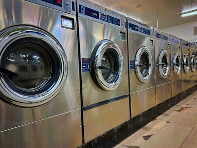 A row of industrial washing machines is lined up in a laundromat. The machines are metallic with glass doors, reflecting light from overhead fluorescent lighting. The floor consists of large tiles with visible wear.