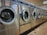 A row of industrial washing machines is lined up in a laundromat. The machines are metallic with glass doors, reflecting light from overhead fluorescent lighting. The floor consists of large tiles with visible wear.