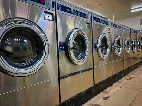 A row of industrial washing machines is lined up in a laundromat. The machines are metallic with glass doors, reflecting light from overhead fluorescent lighting. The floor consists of large tiles with visible wear.