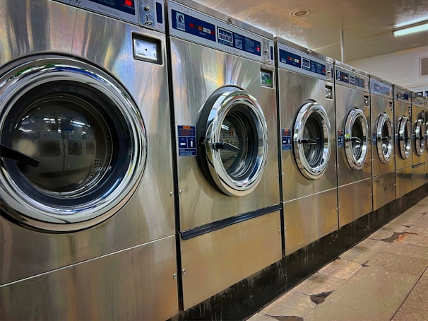 A row of industrial washing machines is lined up in a laundromat. The machines are metallic with glass doors, reflecting light from overhead fluorescent lighting. The floor consists of large tiles with visible wear.