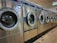 A row of industrial washing machines is lined up in a laundromat. The machines are metallic with glass doors, reflecting light from overhead fluorescent lighting. The floor consists of large tiles with visible wear.