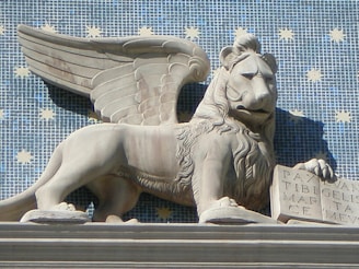 A stone sculpture of a winged lion holding an open book is set against a blue tiled background with yellow star patterns. The lion has detailed features, with a bushy mane and an expression that conveys majesty and strength. The book contains inscriptions and the lion's pose is relaxed yet regal.