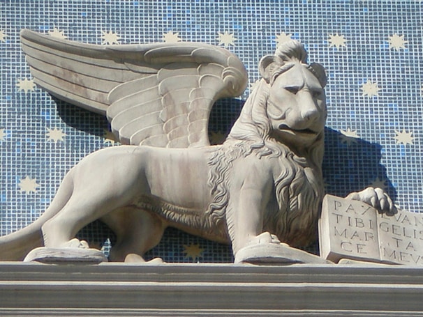 A stone sculpture of a winged lion holding an open book is set against a blue tiled background with yellow star patterns. The lion has detailed features, with a bushy mane and an expression that conveys majesty and strength. The book contains inscriptions and the lion's pose is relaxed yet regal.