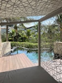 Wooden deck beside a tranquil pool surrounded by lush greenery and palm trees, with modern buildings visible in the background. A geometric-patterned canopy provides partial shade over a seating area.