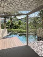 Wooden deck beside a tranquil pool surrounded by lush greenery and palm trees, with modern buildings visible in the background. A geometric-patterned canopy provides partial shade over a seating area.