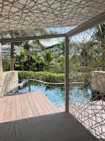 Wooden deck beside a tranquil pool surrounded by lush greenery and palm trees, with modern buildings visible in the background. A geometric-patterned canopy provides partial shade over a seating area.