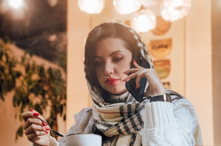 A woman wearing a headscarf is holding a phone to her ear while stirring a drink with a spoon. She is seated indoors, with a hint of greenery in the background and warm, glowing lights above. She is wearing a white sweater and has red lipstick and nail polish.