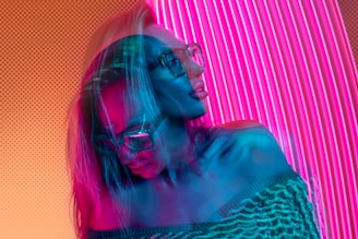 A double-exposed effect creates a dynamic and surreal impression with vibrant colors. A person with long hair wears glasses and a textured off-shoulder sweater. The backdrop consists of vertical pink neon lights, and the composition evokes a sense of futuristic, digital art.