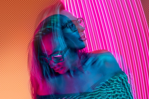 A double-exposed effect creates a dynamic and surreal impression with vibrant colors. A person with long hair wears glasses and a textured off-shoulder sweater. The backdrop consists of vertical pink neon lights, and the composition evokes a sense of futuristic, digital art.