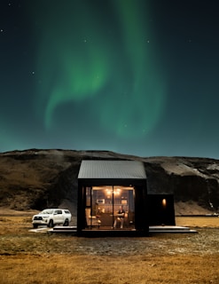 A modern cabin with large glass windows is situated in a remote landscape under the green glow of the Northern Lights. A person is visible inside, sitting at a table, surrounded by a cozy, warmly lit interior. Outside, a white SUV is parked on the grassy terrain near rocky hills.