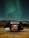 A modern cabin with large glass windows is situated in a remote landscape under the green glow of the Northern Lights. A person is visible inside, sitting at a table, surrounded by a cozy, warmly lit interior. Outside, a white SUV is parked on the grassy terrain near rocky hills.