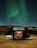 A modern cabin with large glass windows is situated in a remote landscape under the green glow of the Northern Lights. A person is visible inside, sitting at a table, surrounded by a cozy, warmly lit interior. Outside, a white SUV is parked on the grassy terrain near rocky hills.