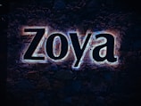 Large illuminated letters spelling 'Zoya' are mounted on a stone wall, with a glow effect highlighting the contours of each letter. The background consists of dark, natural stones.