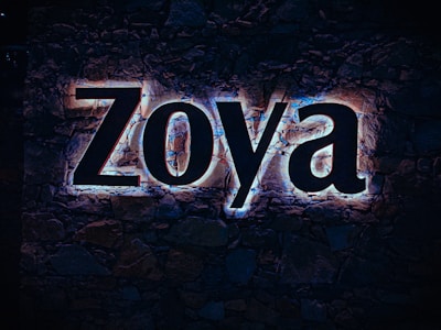 Large illuminated letters spelling 'Zoya' are mounted on a stone wall, with a glow effect highlighting the contours of each letter. The background consists of dark, natural stones.