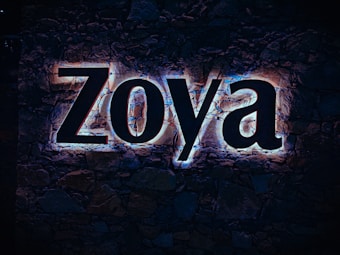 Large illuminated letters spelling 'Zoya' are mounted on a stone wall, with a glow effect highlighting the contours of each letter. The background consists of dark, natural stones.