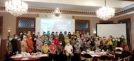 A large group of people are gathered in a conference room for a 'Sensitivity Training' session. Most attendees are wearing face masks. The room is decorated with chandeliers and a large screen displaying the title of the seminar. Attendees are dressed in various colorful garments.